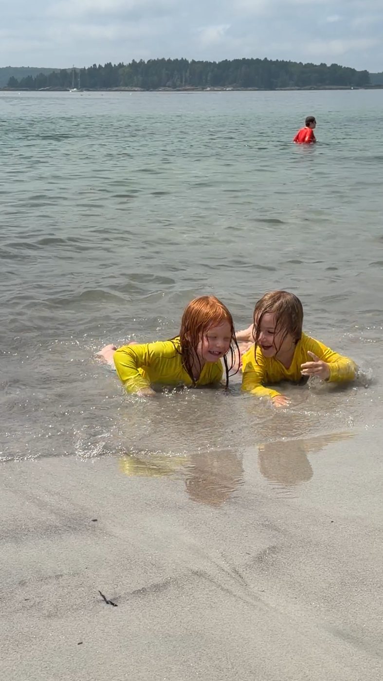 Two children lie laughing in the surf at a Maine beach